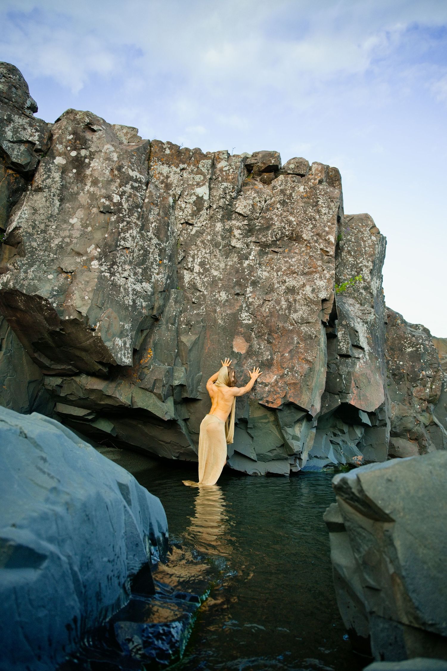 Woman in sheer dress stands in water reaching toward massive lichen-covered rock formation.