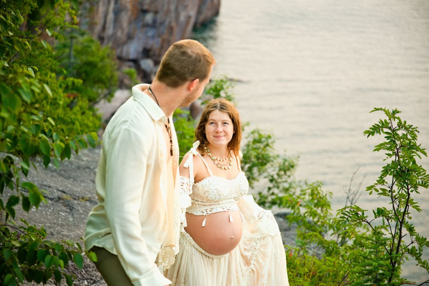 Pregnant couple posing outdoors near a rocky cliff and water, wearing bohemian cream outfits during a maternity photo shoot.