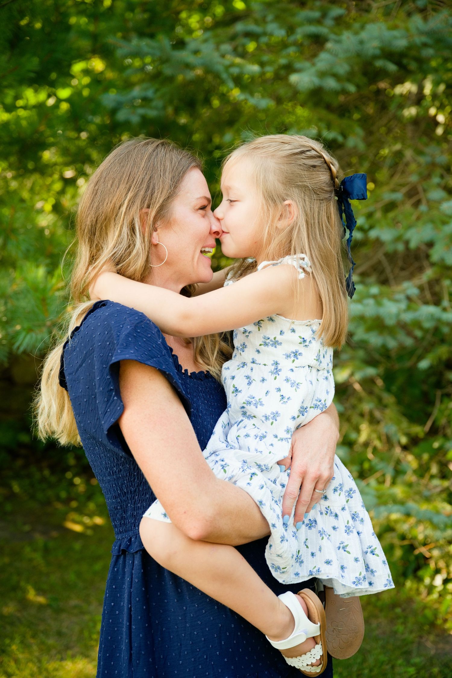 A mother in a navy dress holds her young blonde daughter in a floral dress, sharing an eskimo kiss outdoors.