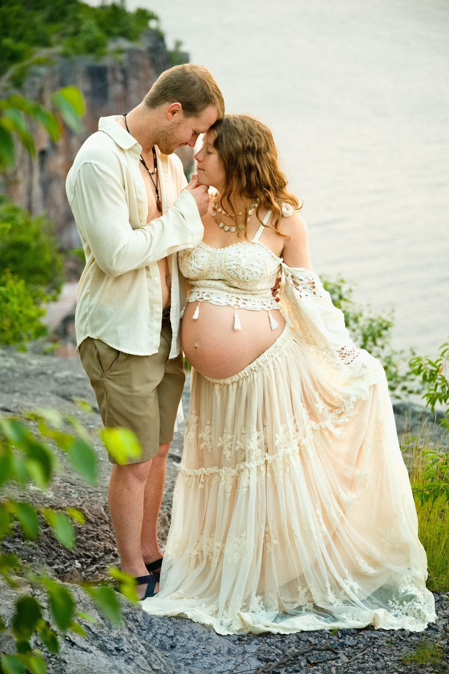 Couple sharing a tender moment outdoors, pregnant woman in lace dress, man in casual attire, rocky waterside backdrop.