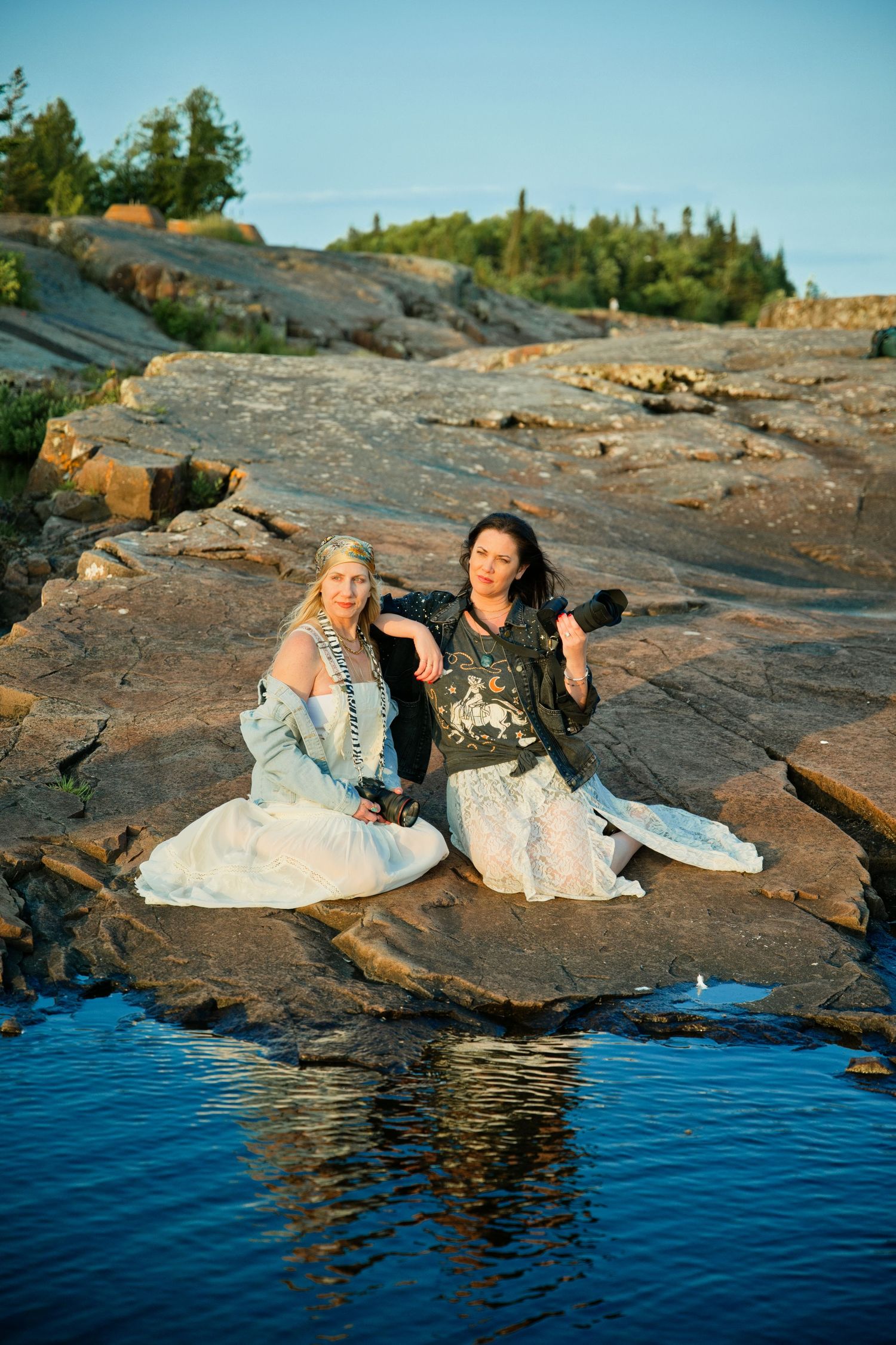 Two women in elegant dresses sit on rocky shoreline near calm water with trees in background.