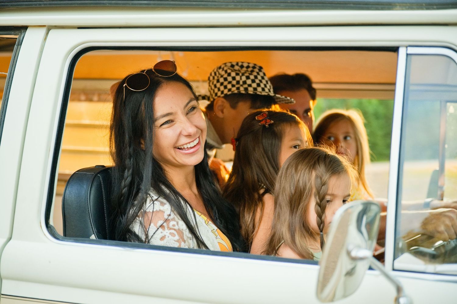 A smiling woman with long dark hair sits in a vintage van window with friends and children during a road trip.