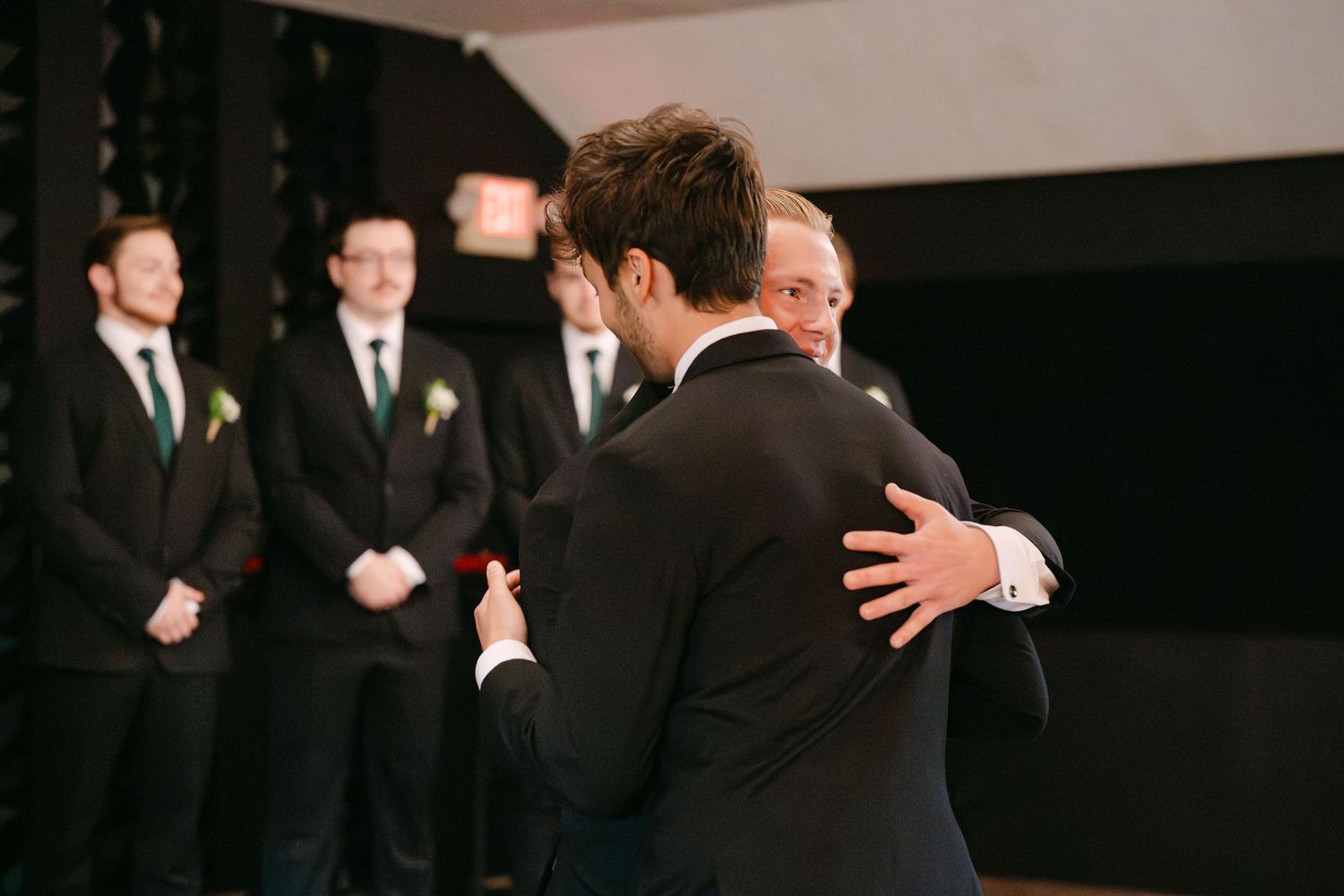 Groomsmen in black suits share an emotional embrace during a wedding ceremony.