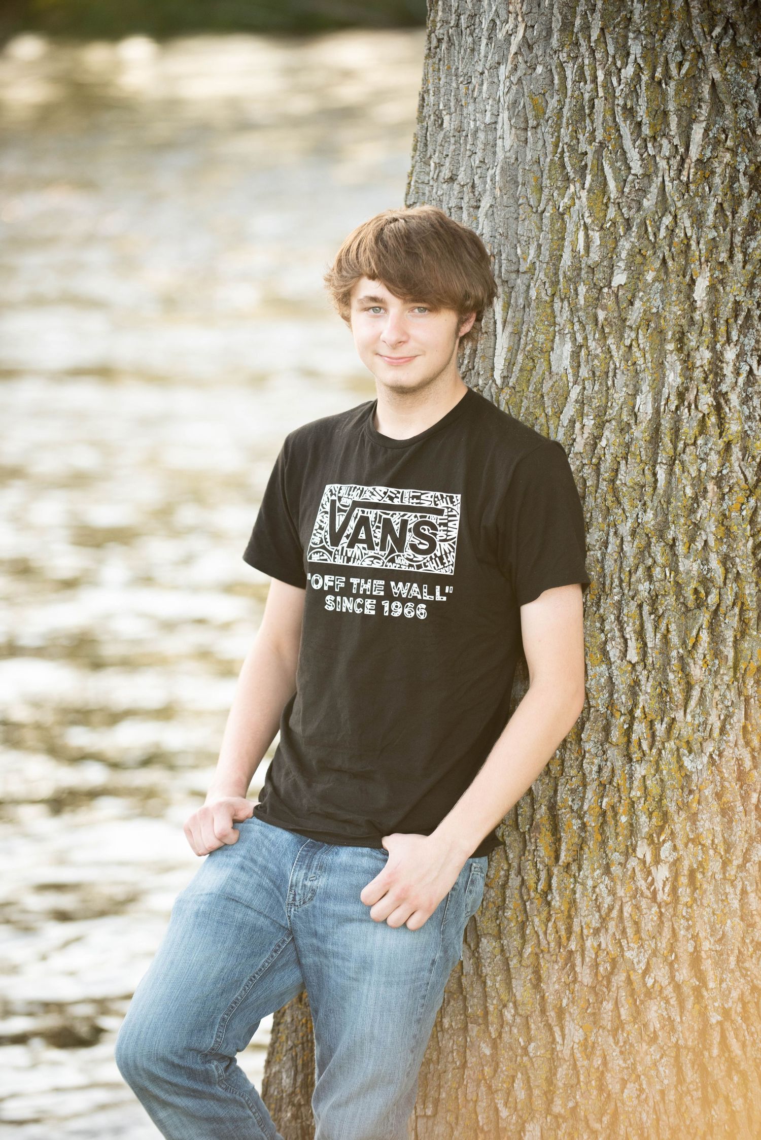 Person wearing Vans t-shirt and jeans leaning against tree trunk near water.