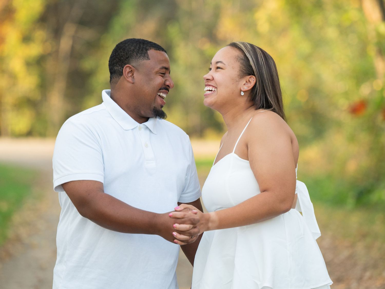 A happy couple laughs and embraces while taking romantic engagement photos in a sunlit outdoor setting.