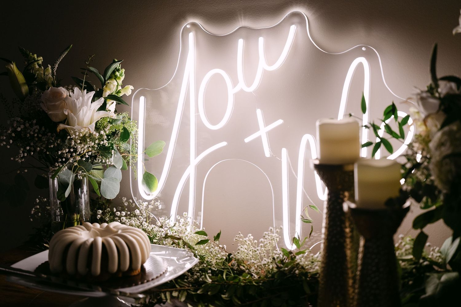 Elegant wedding dessert table with neon sign, bundt cake, candles and floral arrangements.