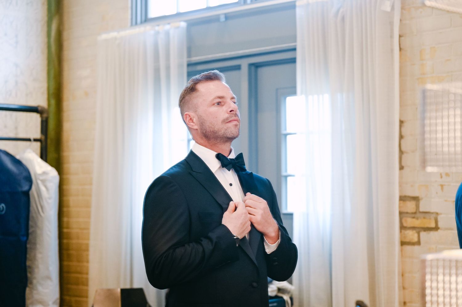 A person in a black suit adjusts their tie while standing in front of sheer white curtains.