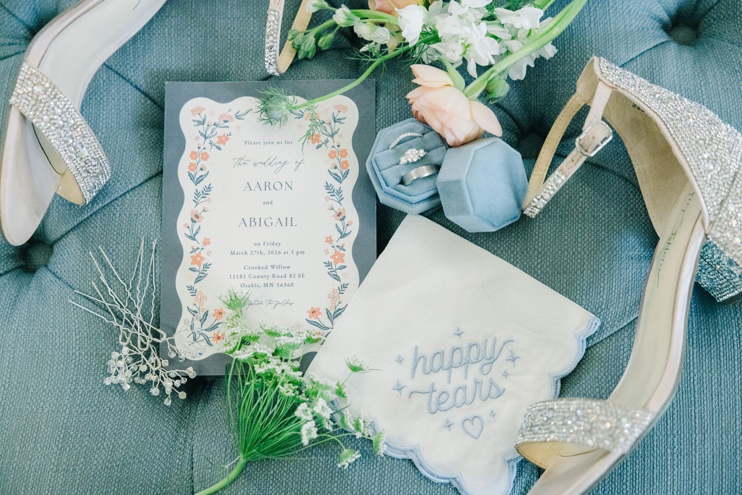Elegant wedding flat lay with glitter heels, floral invitation, ring box, and happy tears handkerchief on blue velvet.