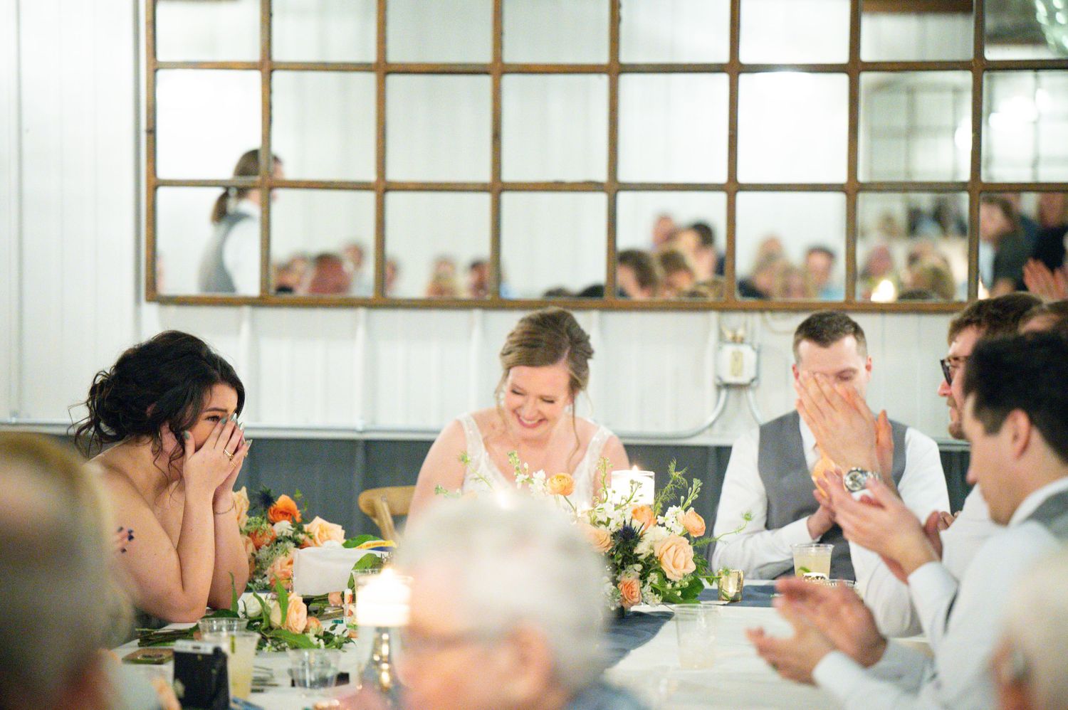 Bride and wedding guests laugh during speeches at floral-decorated head table at wedding reception.