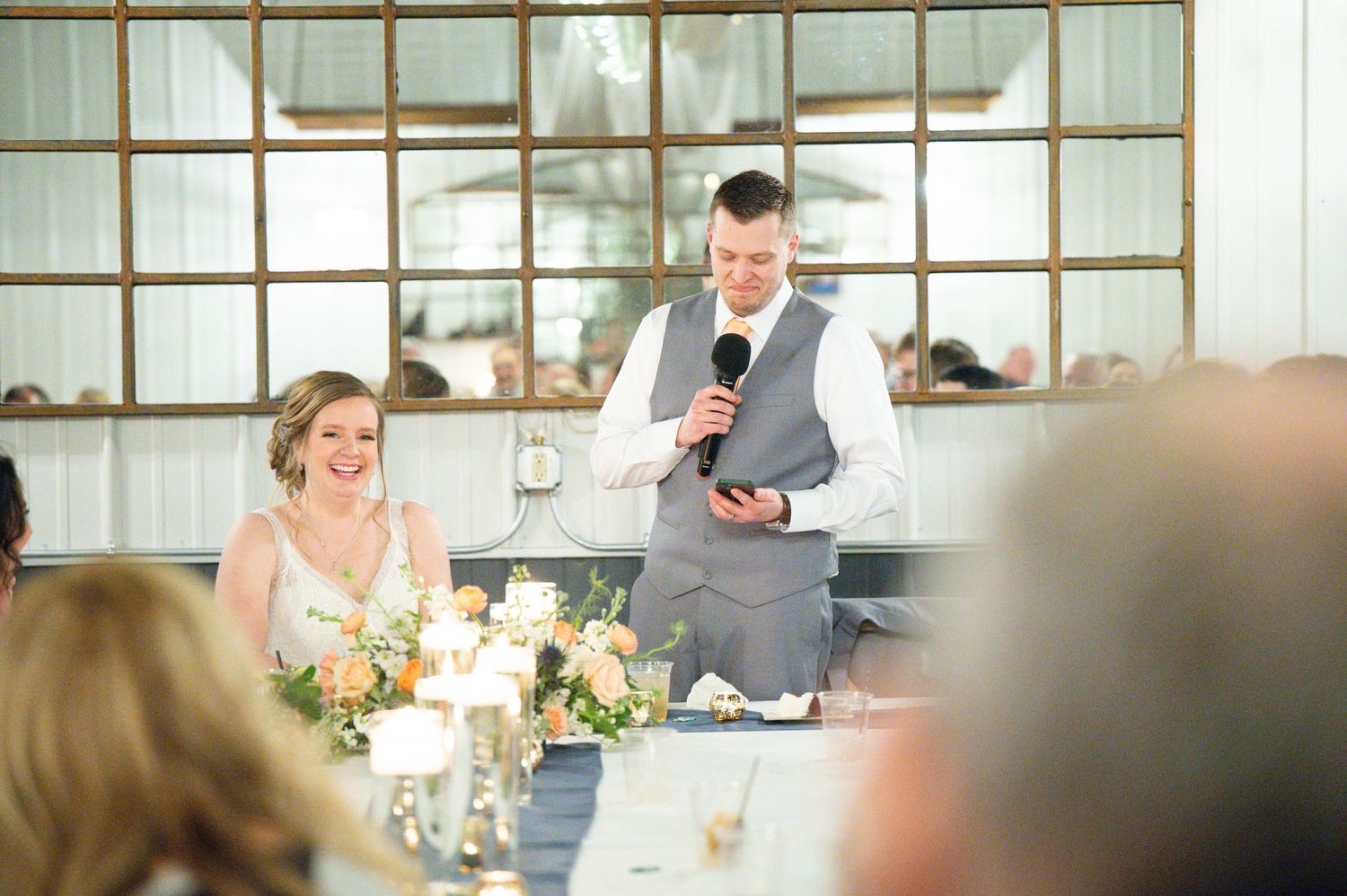 Groom speaks into microphone at wedding reception while bride laughs joyfully at decorated head table.