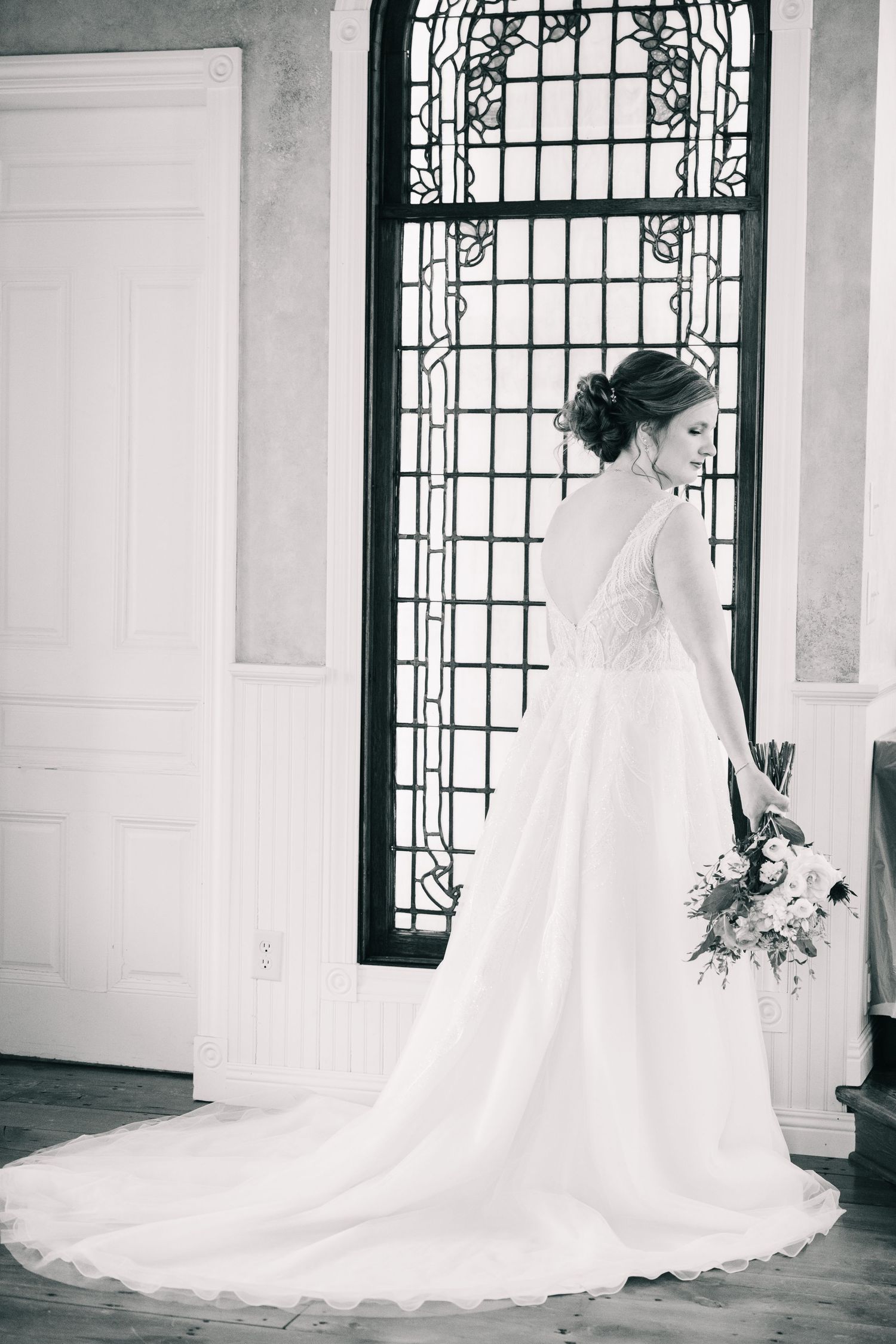 Black and white portrait of bride in flowing white gown with train standing before ornate stained glass window.