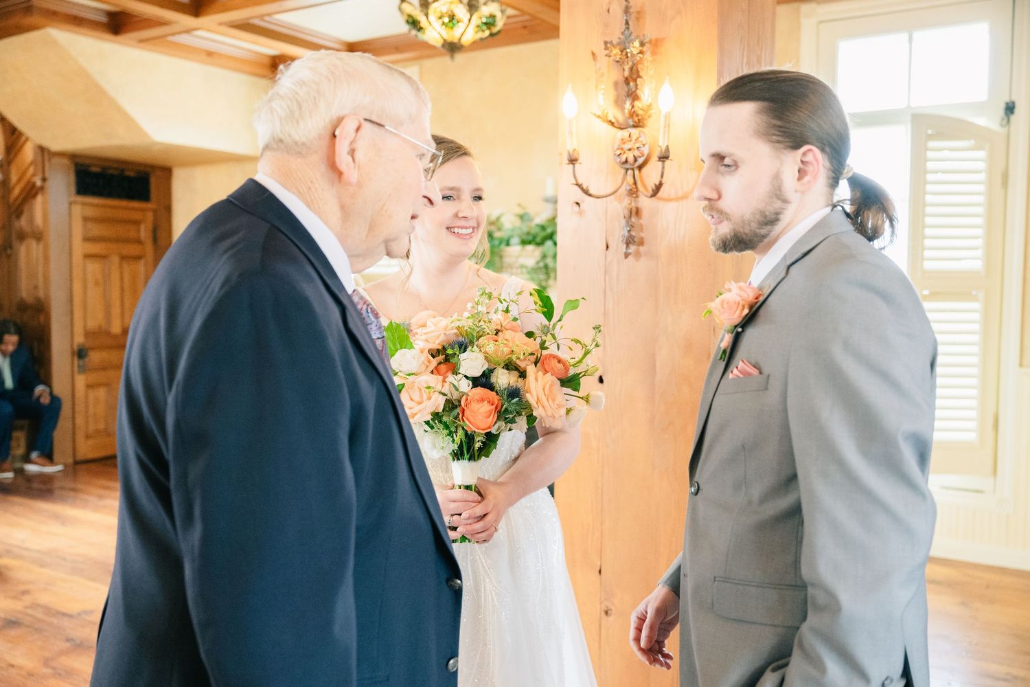 Father of bride presents daughter to groom during intimate indoor wedding ceremony with chandelier.