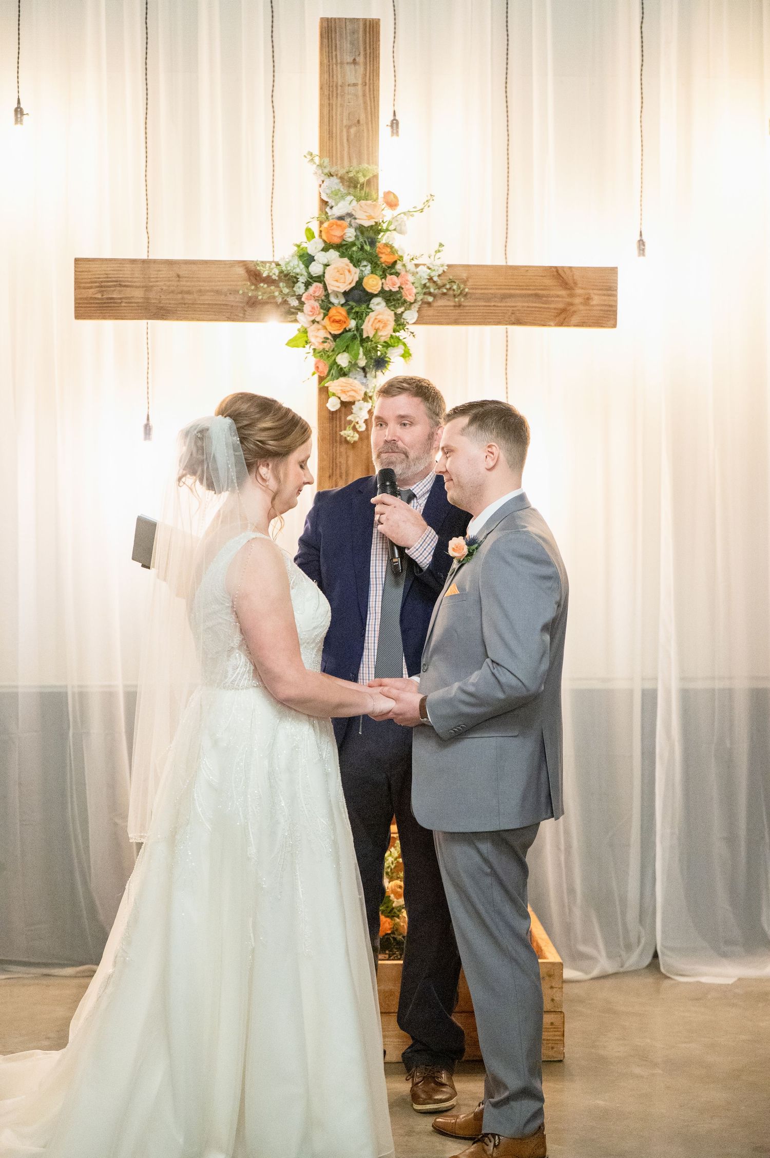 Bride and groom exchange vows before officiant at altar decorated with floral cross and white draping.