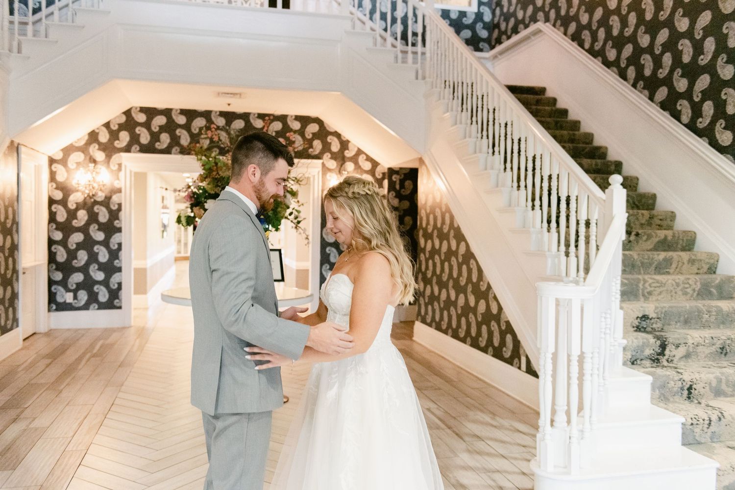 Central MN Wedding Photos | Bride and groom embracing during emotional first look beside a grand staircase with paisley walls.