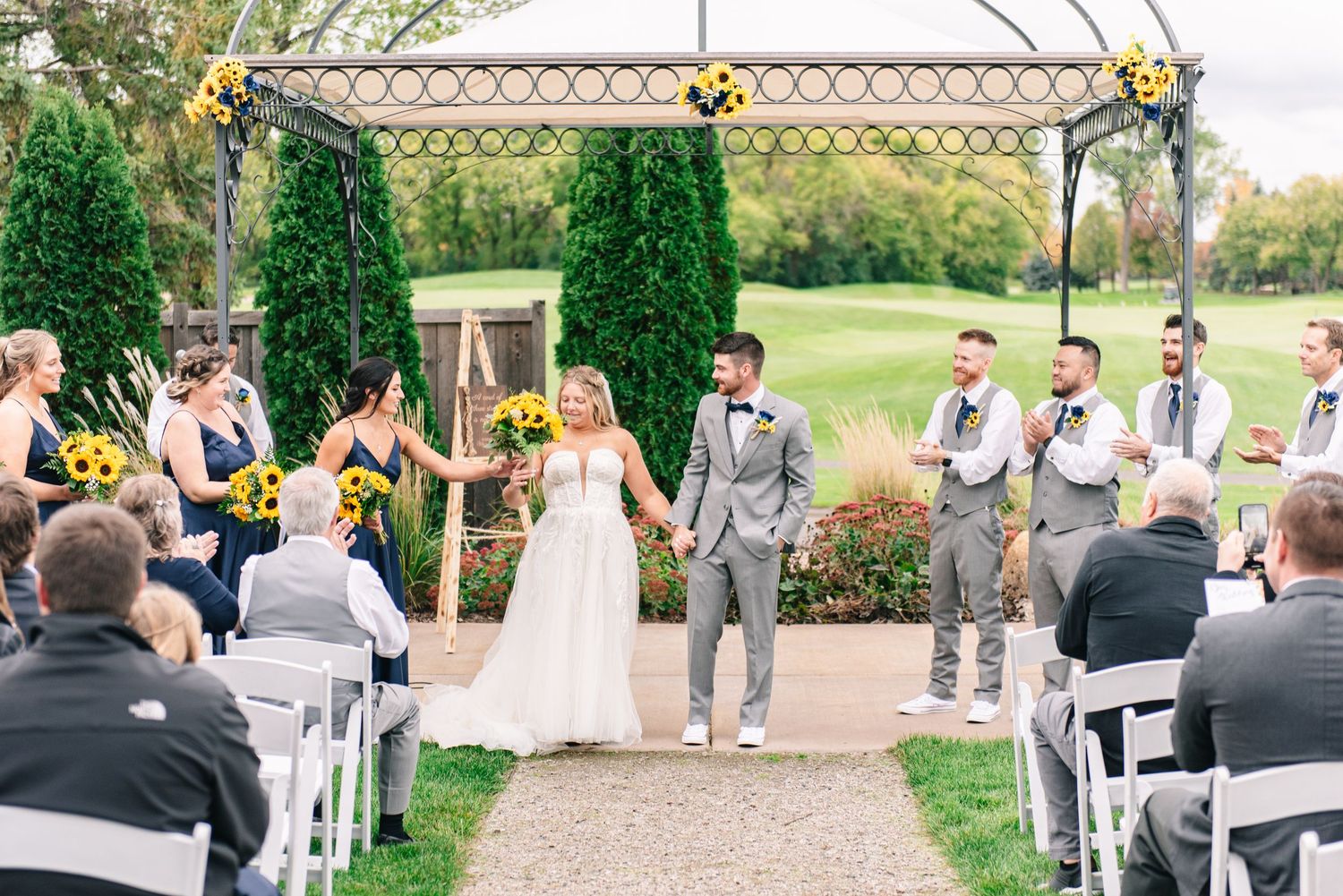 Central MN Wedding Photos - Newlyweds raise hands in celebration at outdoor ceremony altar with groomsmen and sunflower decorations.