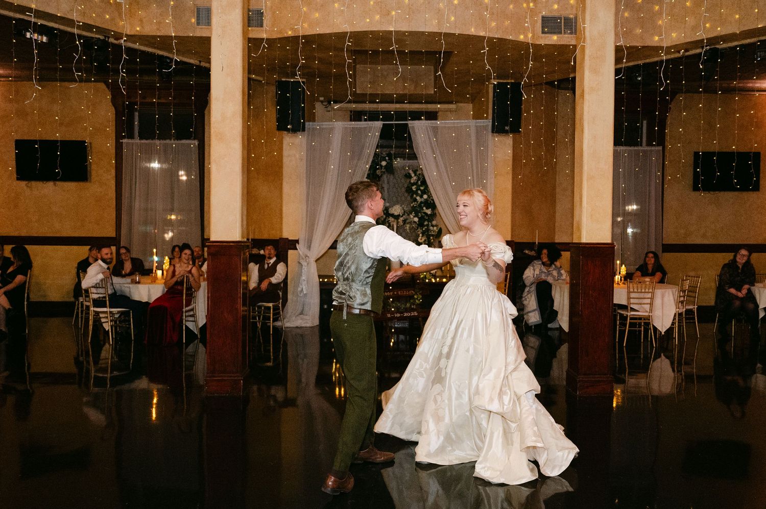 Central MN Wedding Photos | Bride and groom sharing their first dance in a warmly lit ballroom with string lights.