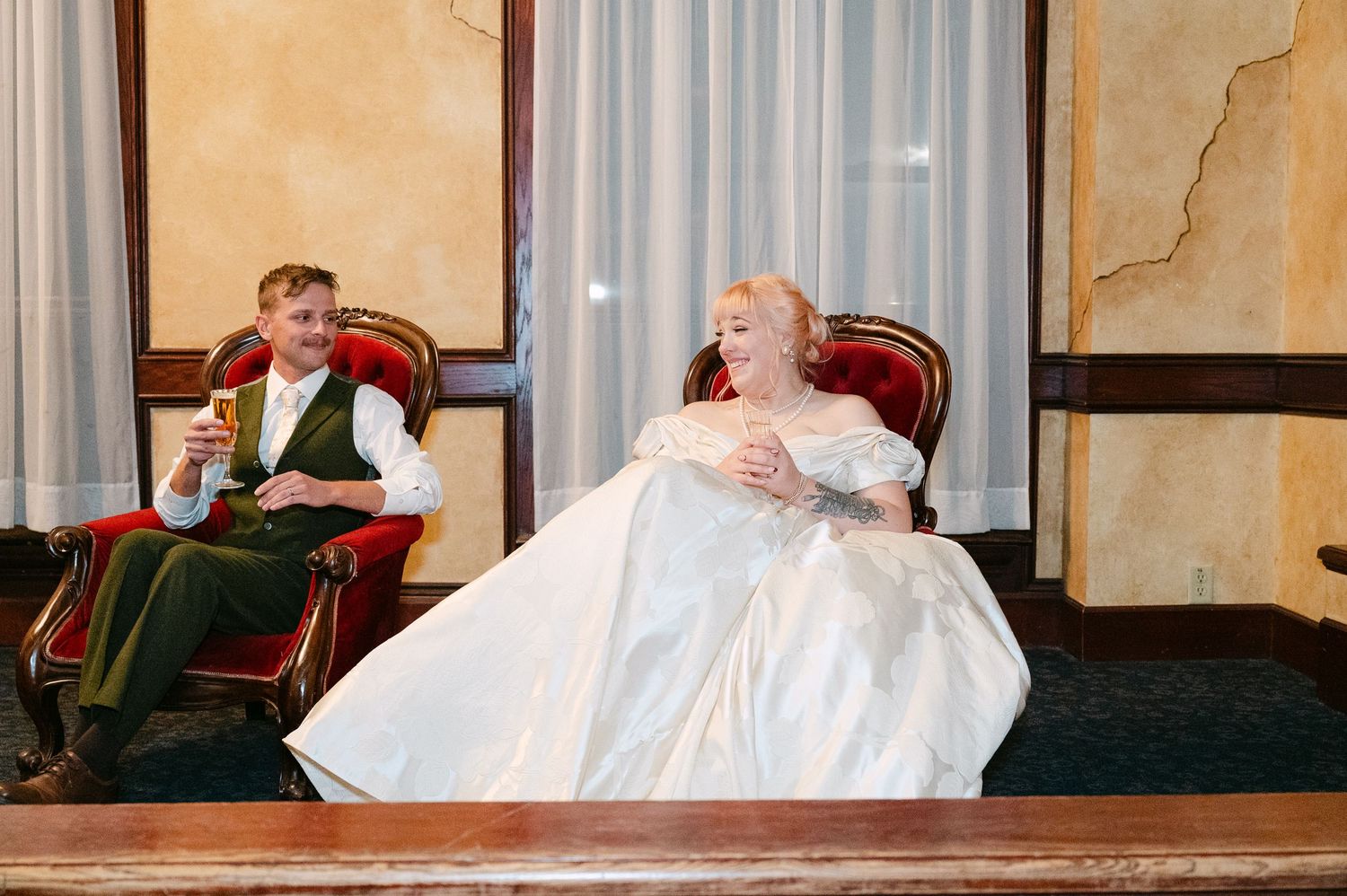Central MN Wedding Photos | Bride and groom relax in vintage red velvet chairs during wedding reception at historic venue.