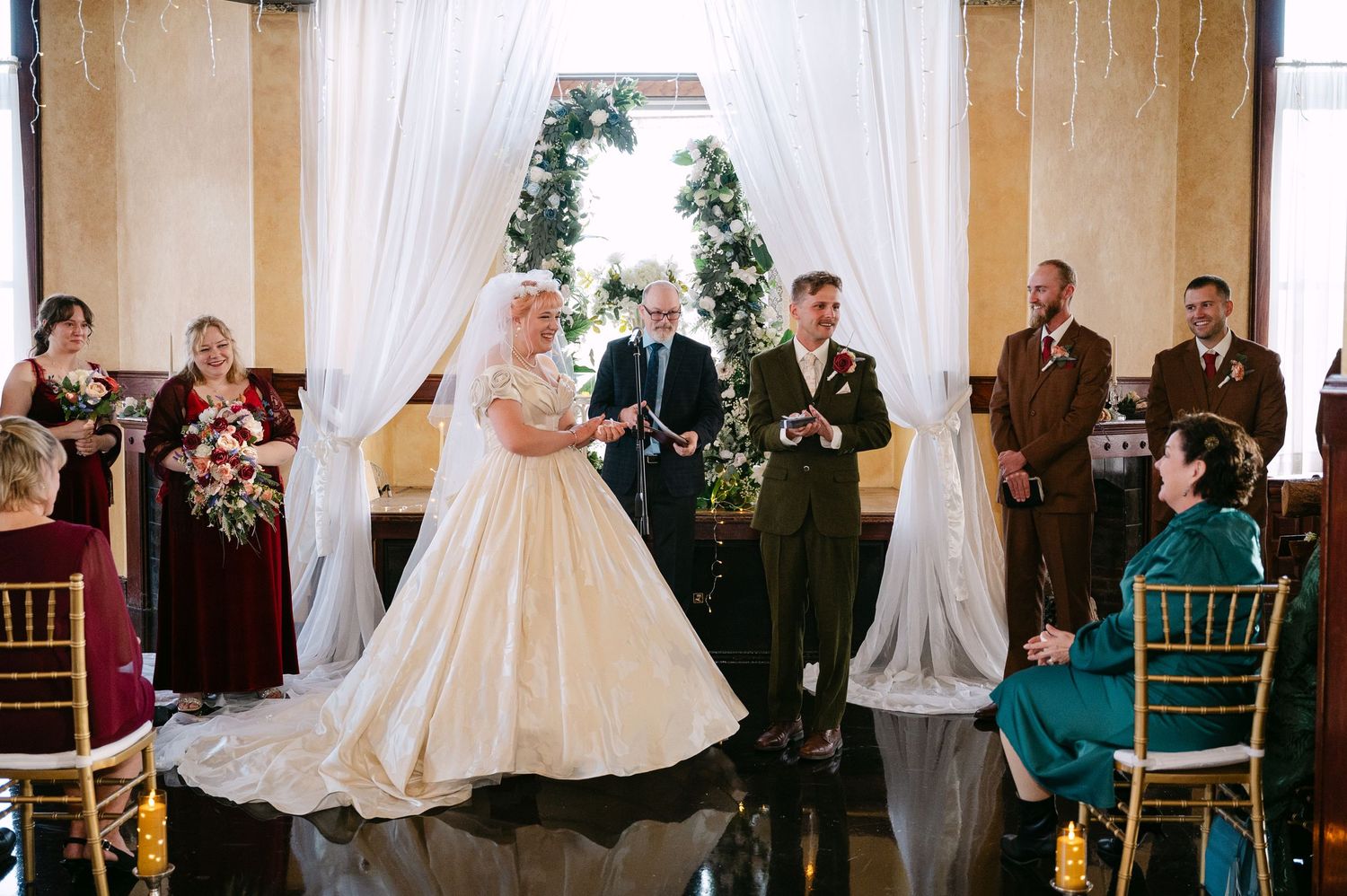 Central MN Wedding Photos | Bride and groom exchange vows at elegant wedding ceremony with white drapes and greenery.