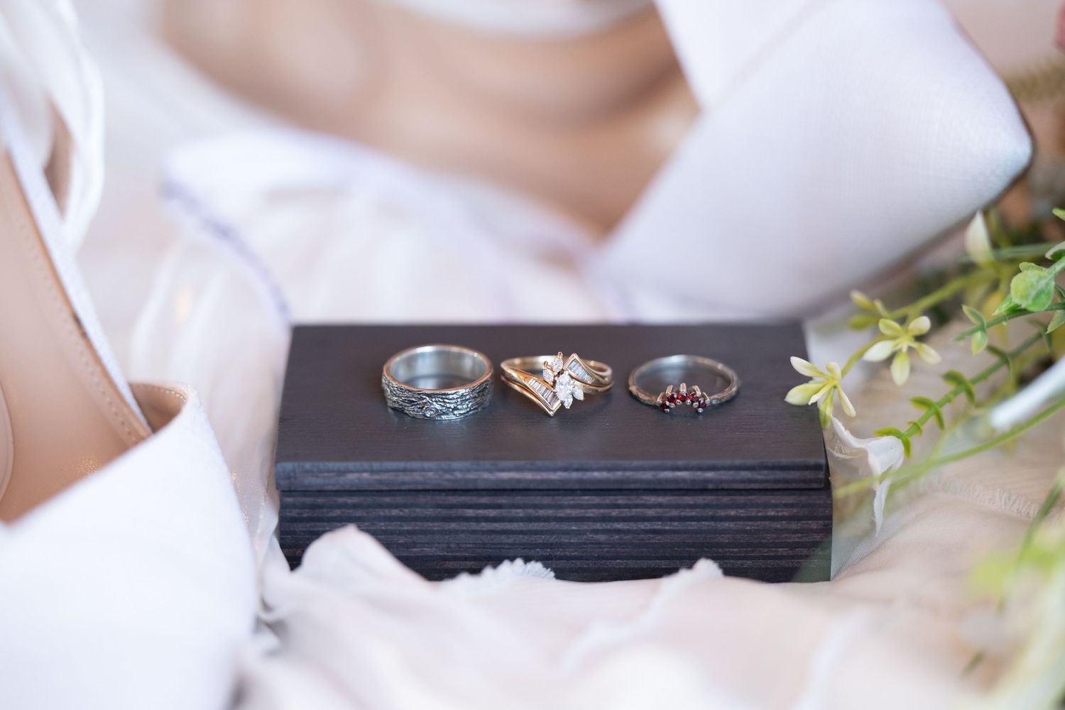 The Historic Concord Exchange 3 elegant wedding rings displayed on a dark jewelry box with white bridal dress and greenery in background.