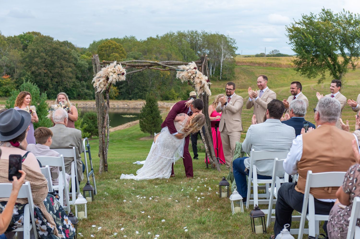 Central MN Wedding Photos - first kiss at outdoor wedding ceremony under floral arch, guests seated on white chairs on green lawn.