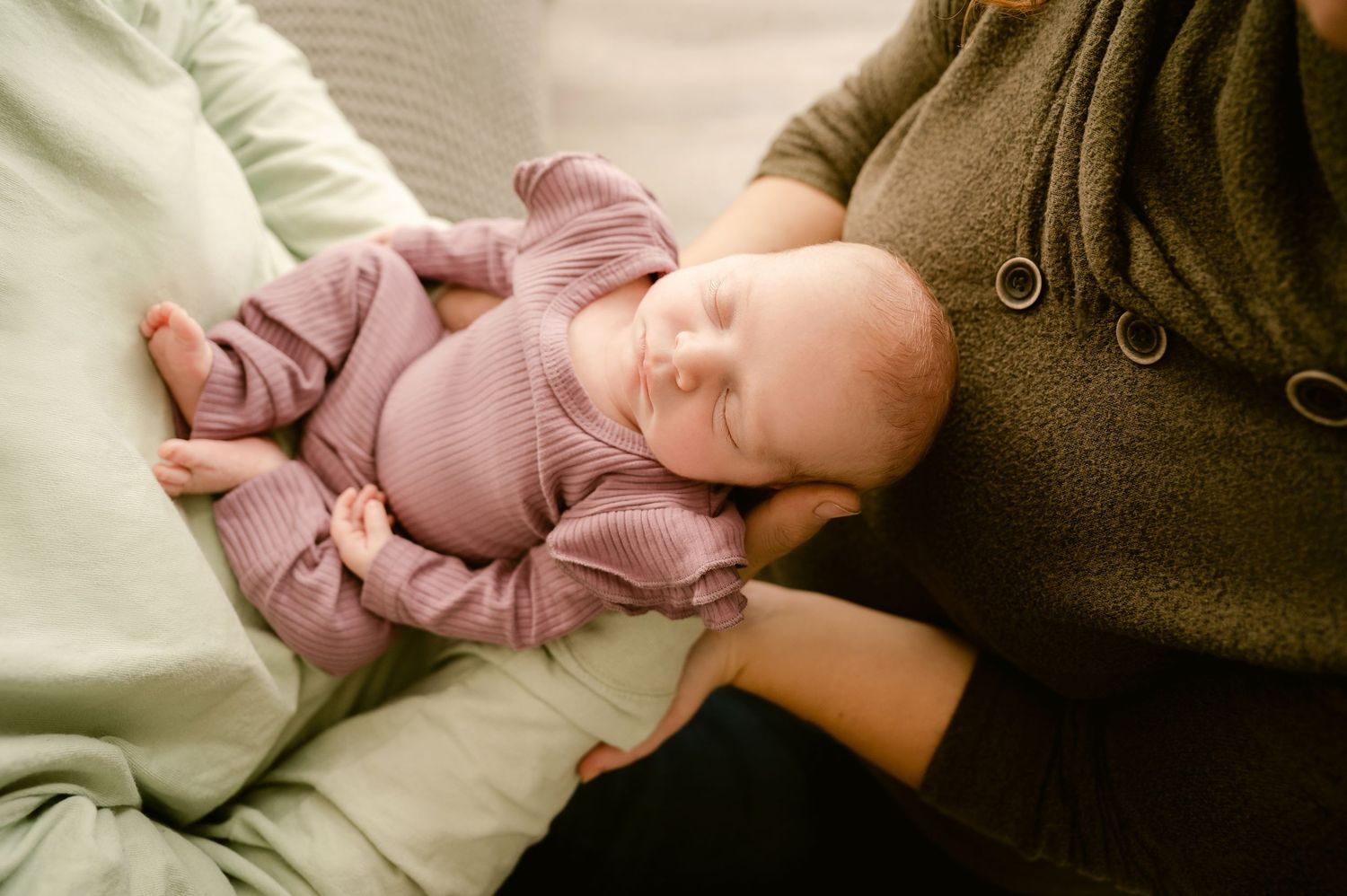 St Cloud newborn Photos of Sleeping newborn in purple knit outfit cradled in parent's arms on soft sofa.