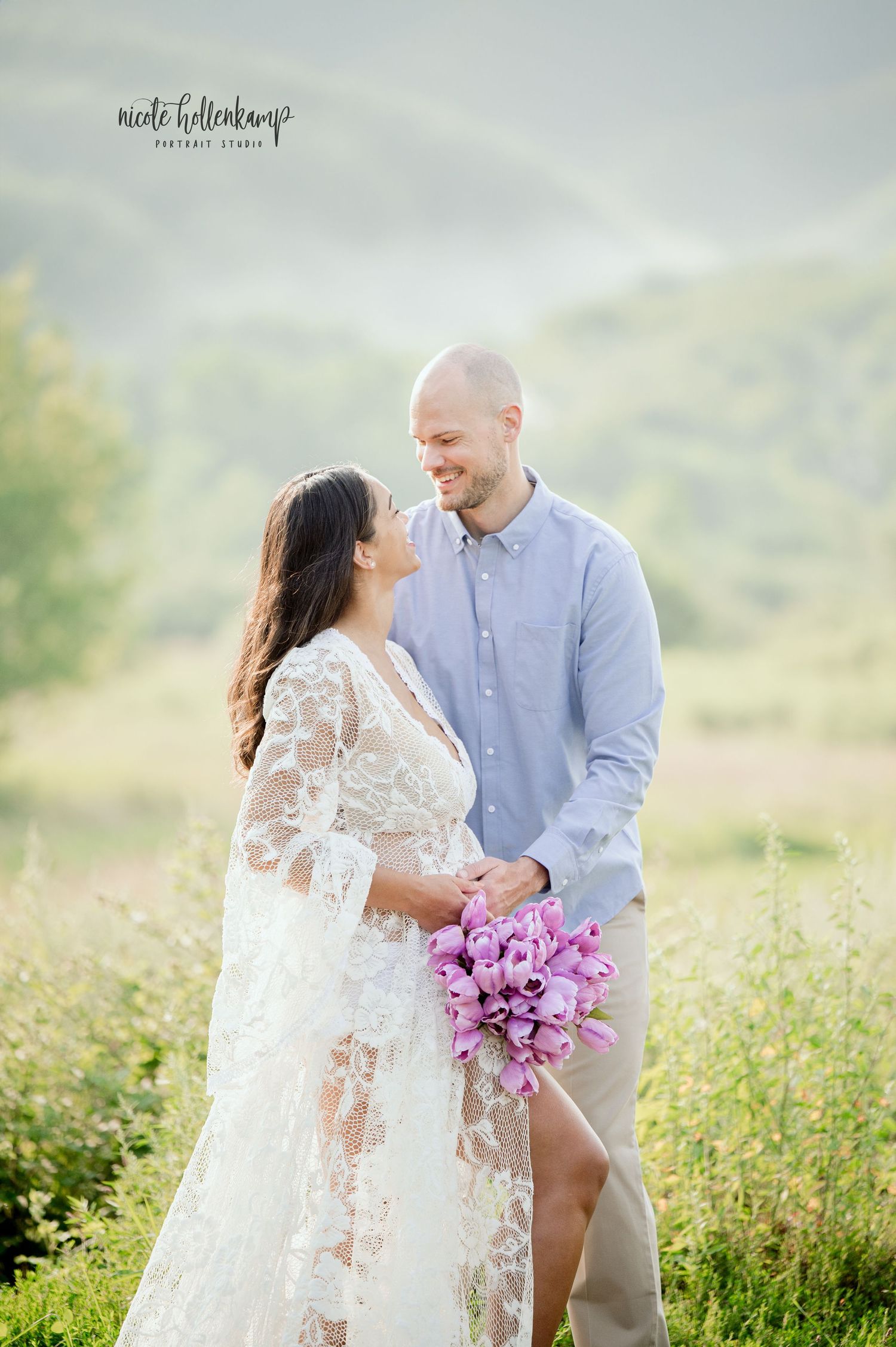 Bride in white lace gown holding purple bouquet smiles with groom in blue shirt in a sunlit meadow.