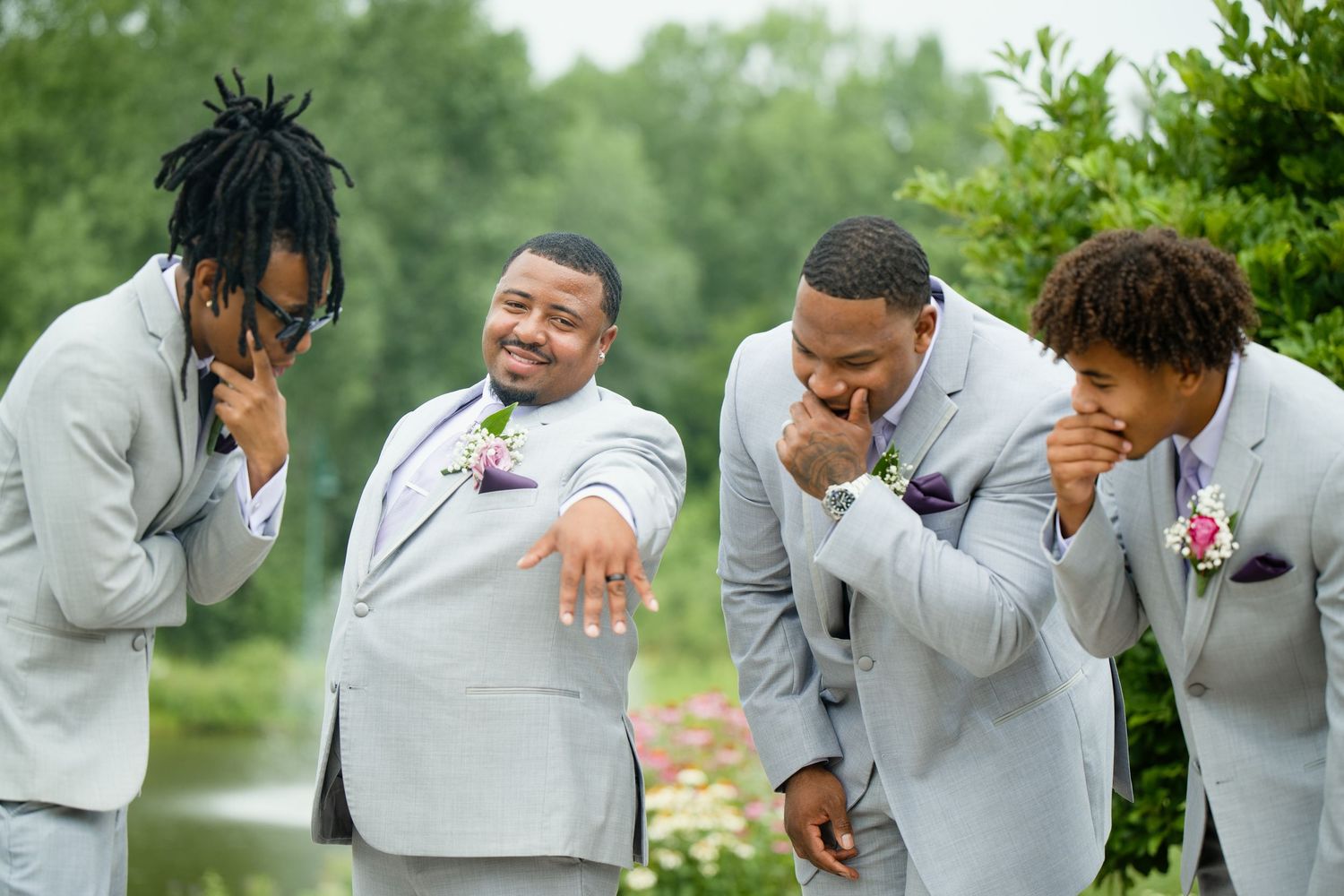 Groom shows off wedding ring to laughing groomsmen in gray suits with purple boutonnieres outdoors.
