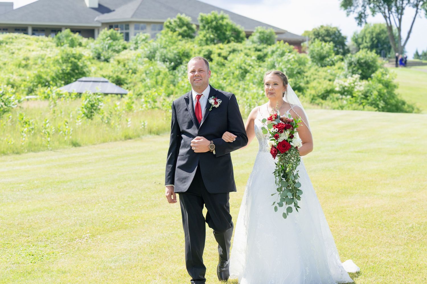Father walks bride in white gown holding red rose bouquet down aisle on sunny wedding day.
