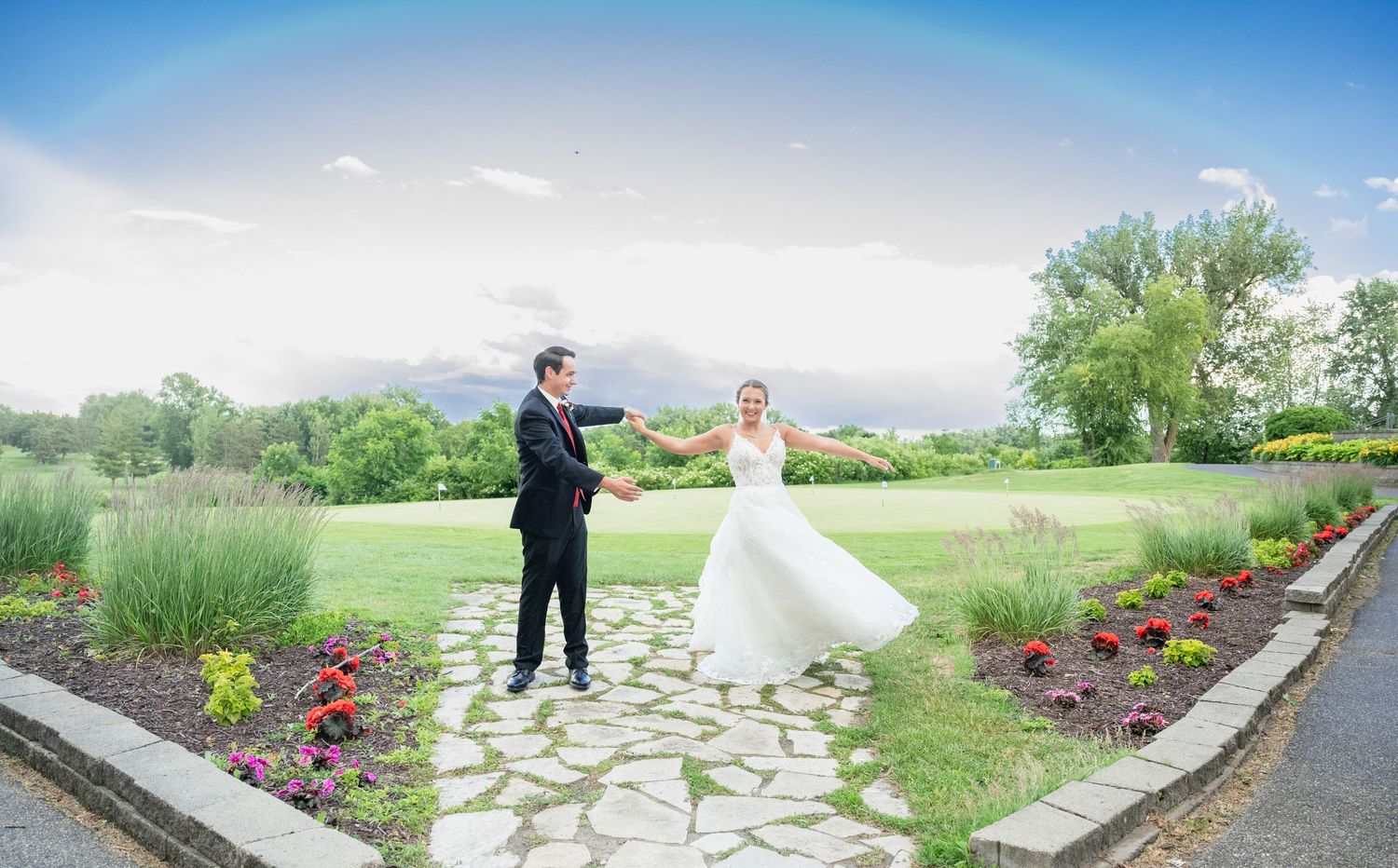Bride and groom dance on a stone garden path with a beautiful rainbow arching overhead.
