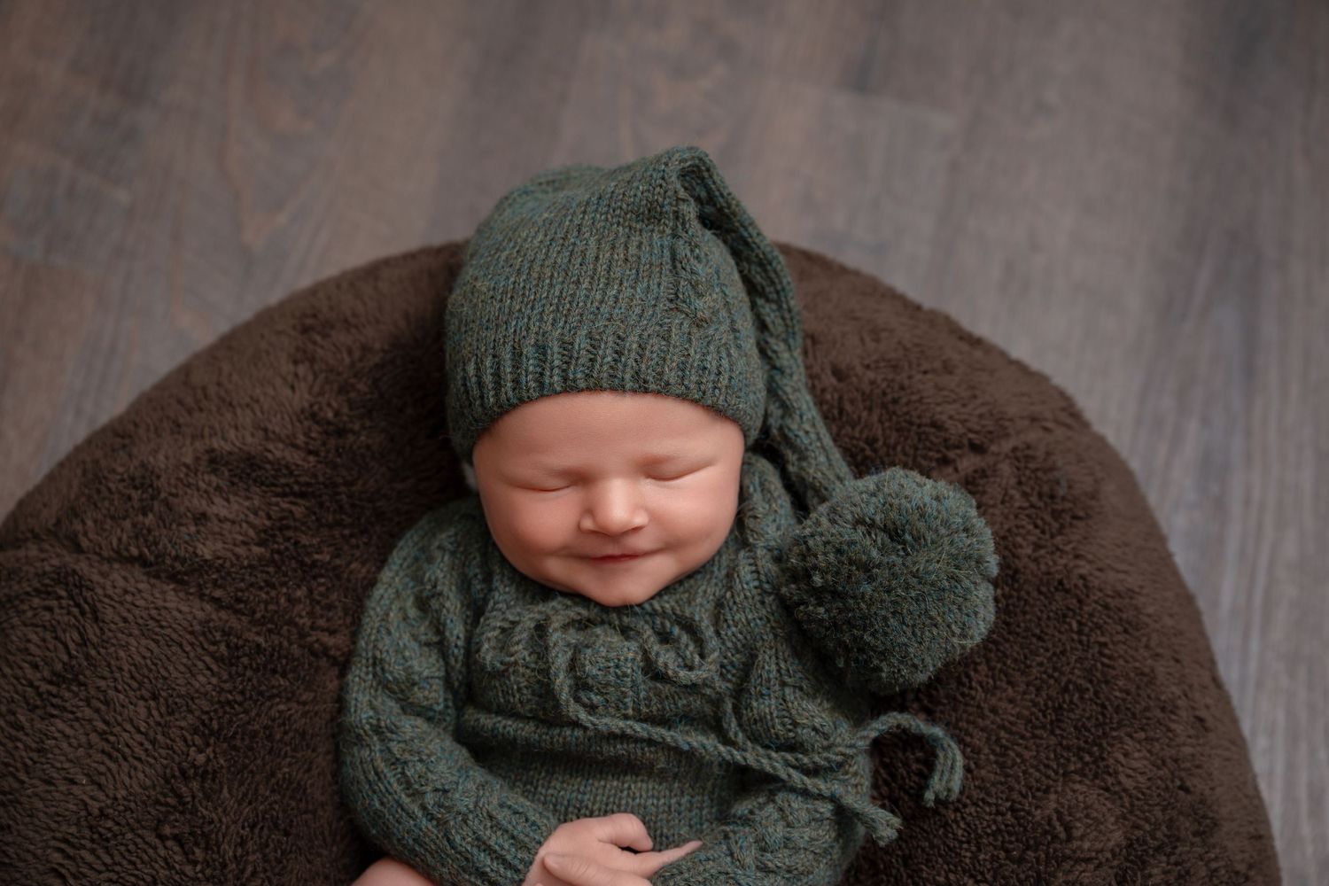 Small baby sleeping peacefully on brown cushion wearing knitted green hat and sweater outfit.