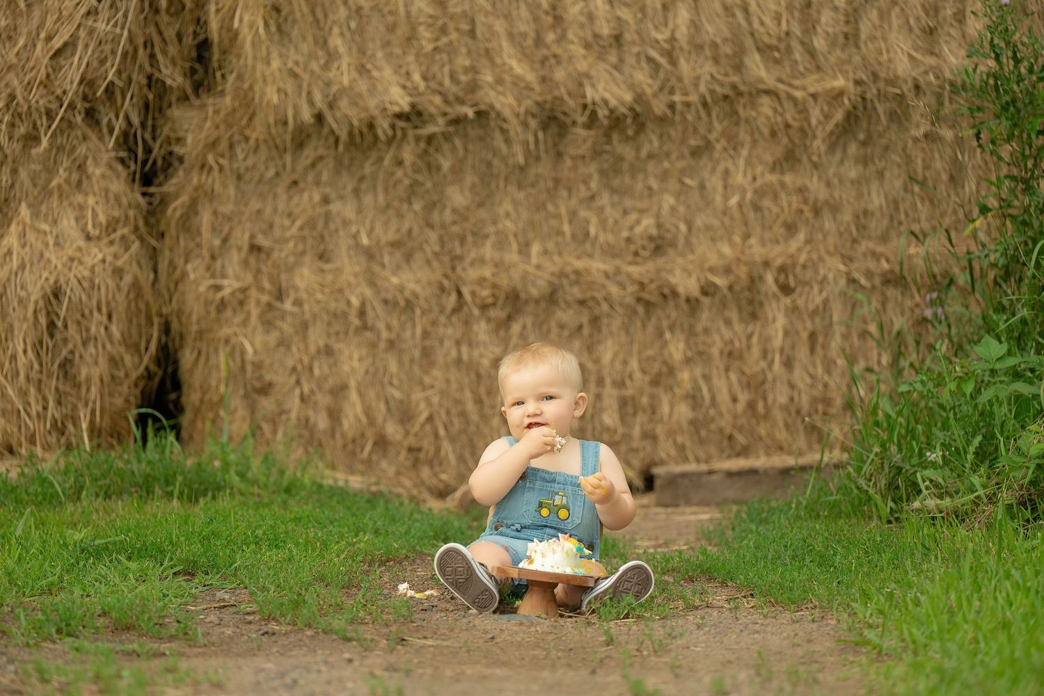 A small child sits on the ground playing near a hay bale wall on a grassy path. Family Portraits & Milestone Moments in Foreston, MN