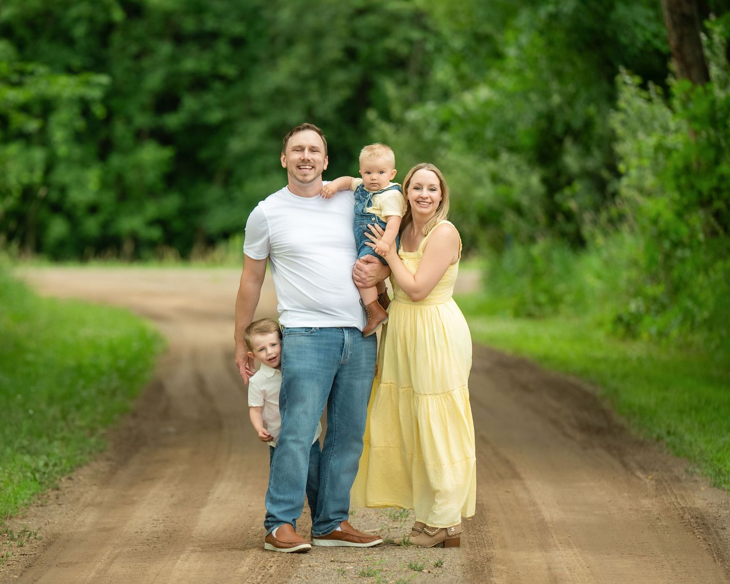 Happy group in a summer photo session walking along a dirt road surrounded by lush green trees and foliage. Family Portraits & Milestone Mom
