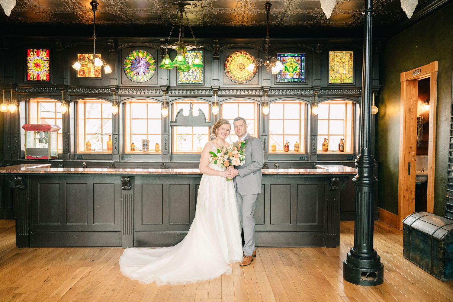 Bride and groom pose together in elegant venue with stained glass windows and dark wood bar backdrop.