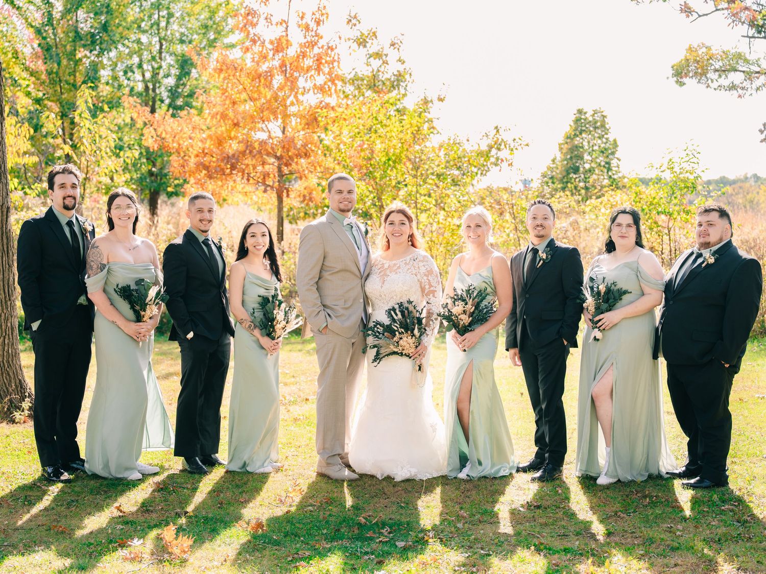 Wedding party in sage green and black formal attire posing outdoors during fall foliage season.