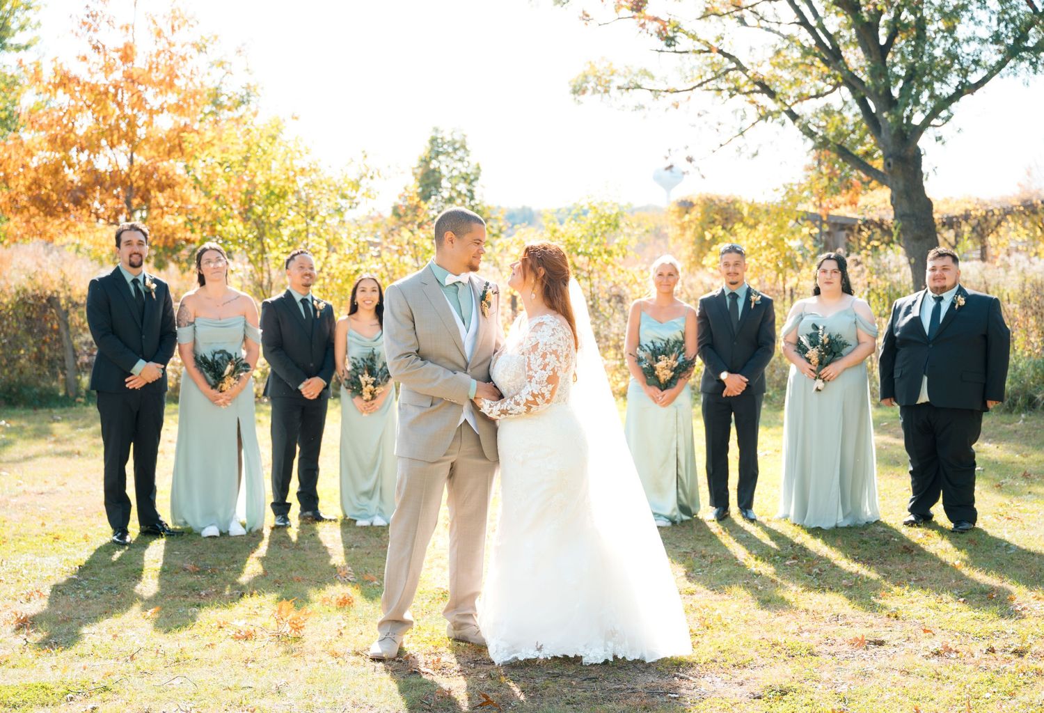 Bride and groom share a moment as wedding party stands behind them in a sunlit autumn field.