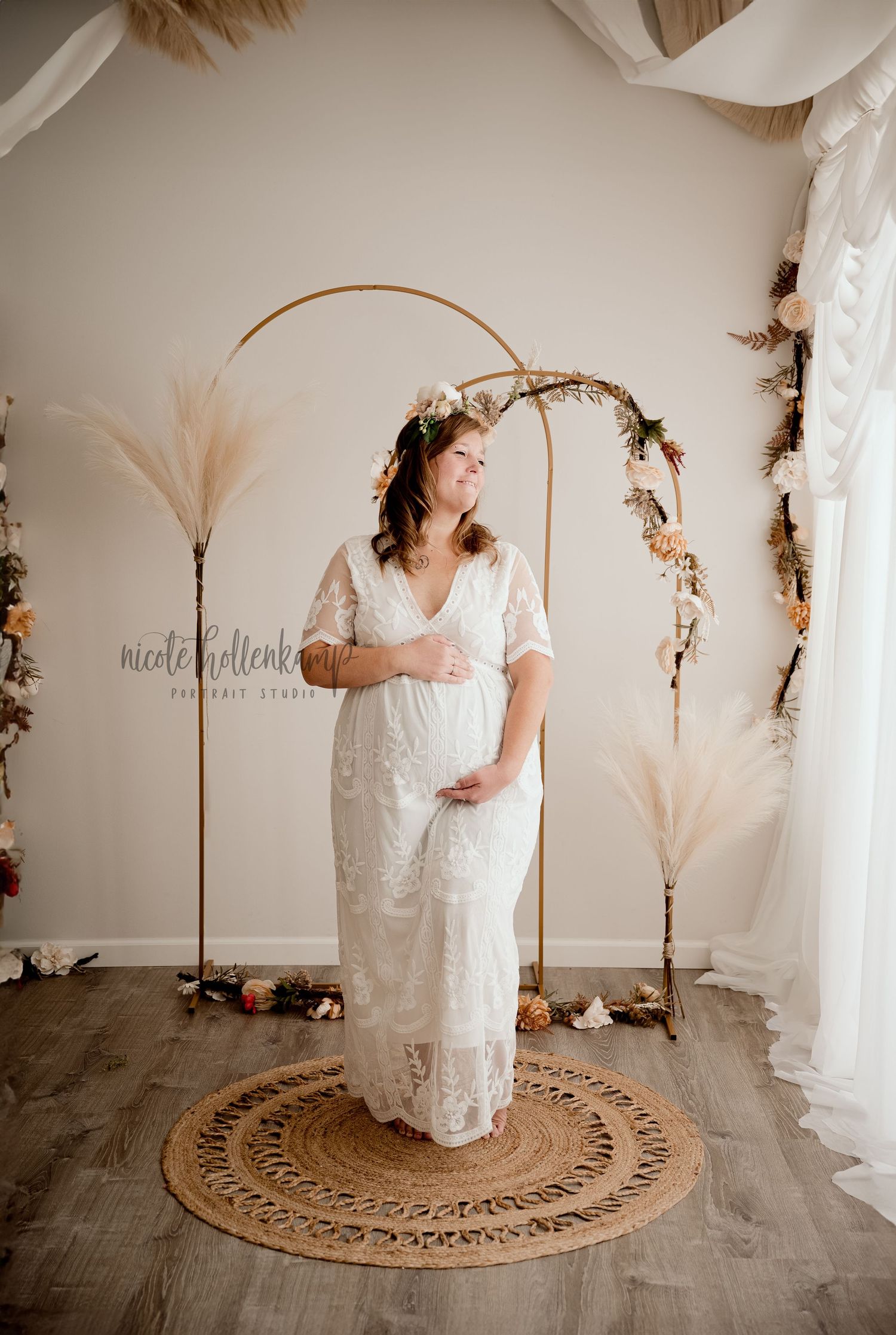 Expectant mother in lace dress poses under boho arch with pampas grass decor.