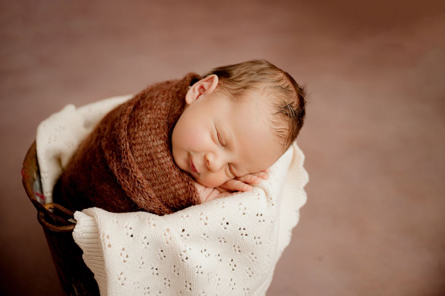 Sleeping newborn with bow accessory wrapped in brown knit blanket on white textured background.