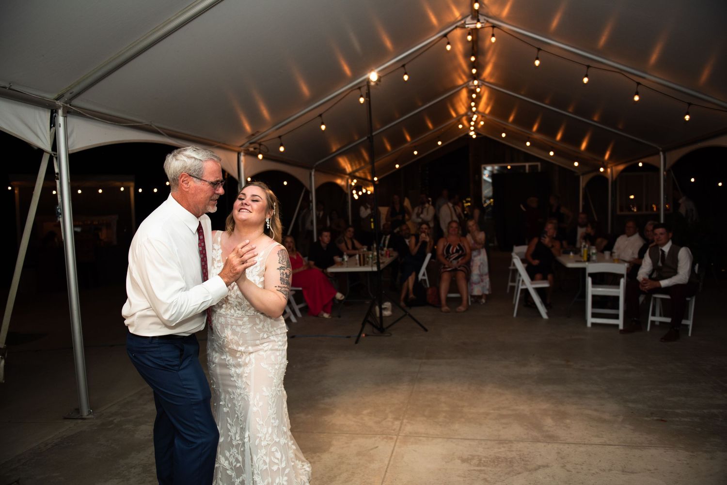Panoramic view of guests dancing under a lit wedding reception tent at night with string lights creating a warm atmosphere.