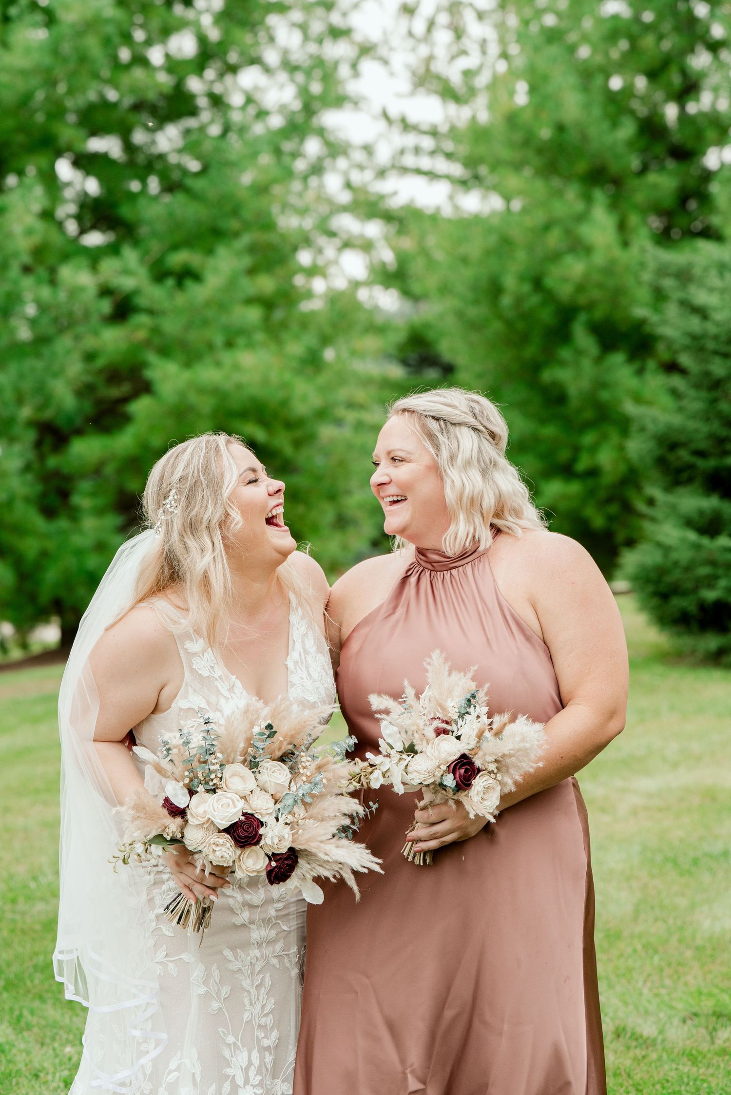 Two women share a joyful moment outdoors while holding elegant white and burgundy floral bouquets.