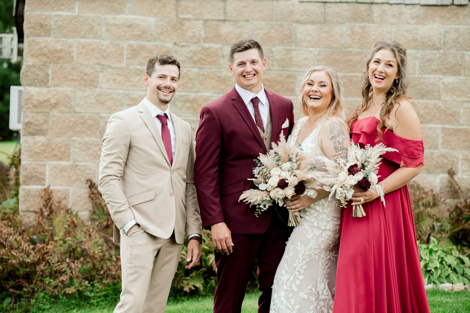 Wedding party members celebrate with laughter in front of stone wall and barn.