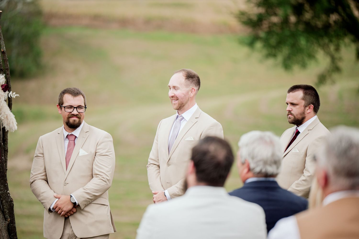 Groomsmen in tan suits stand together during an outdoor wedding ceremony.