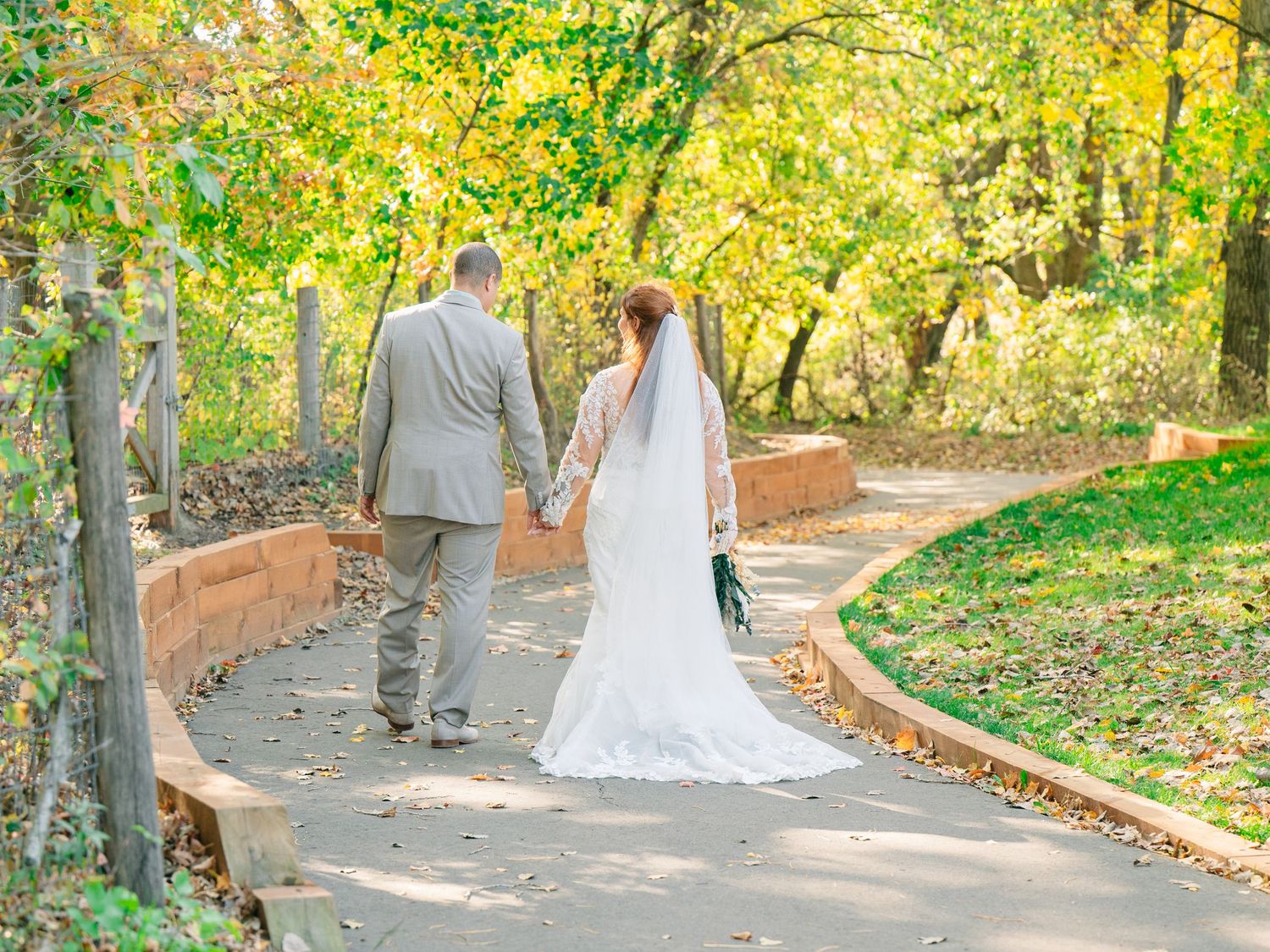 Bride and groom walk hand-in-hand down a tree-lined path during their fall outdoor wedding.