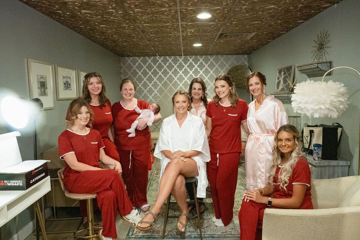 Bridal party in red pajamas and robes smiling with bride holding newborn baby in a salon getting ready room.