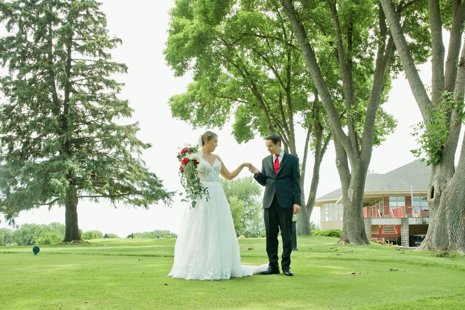 Bride and groom walk hand in hand on green golf course lawn surrounded by tall shady trees.