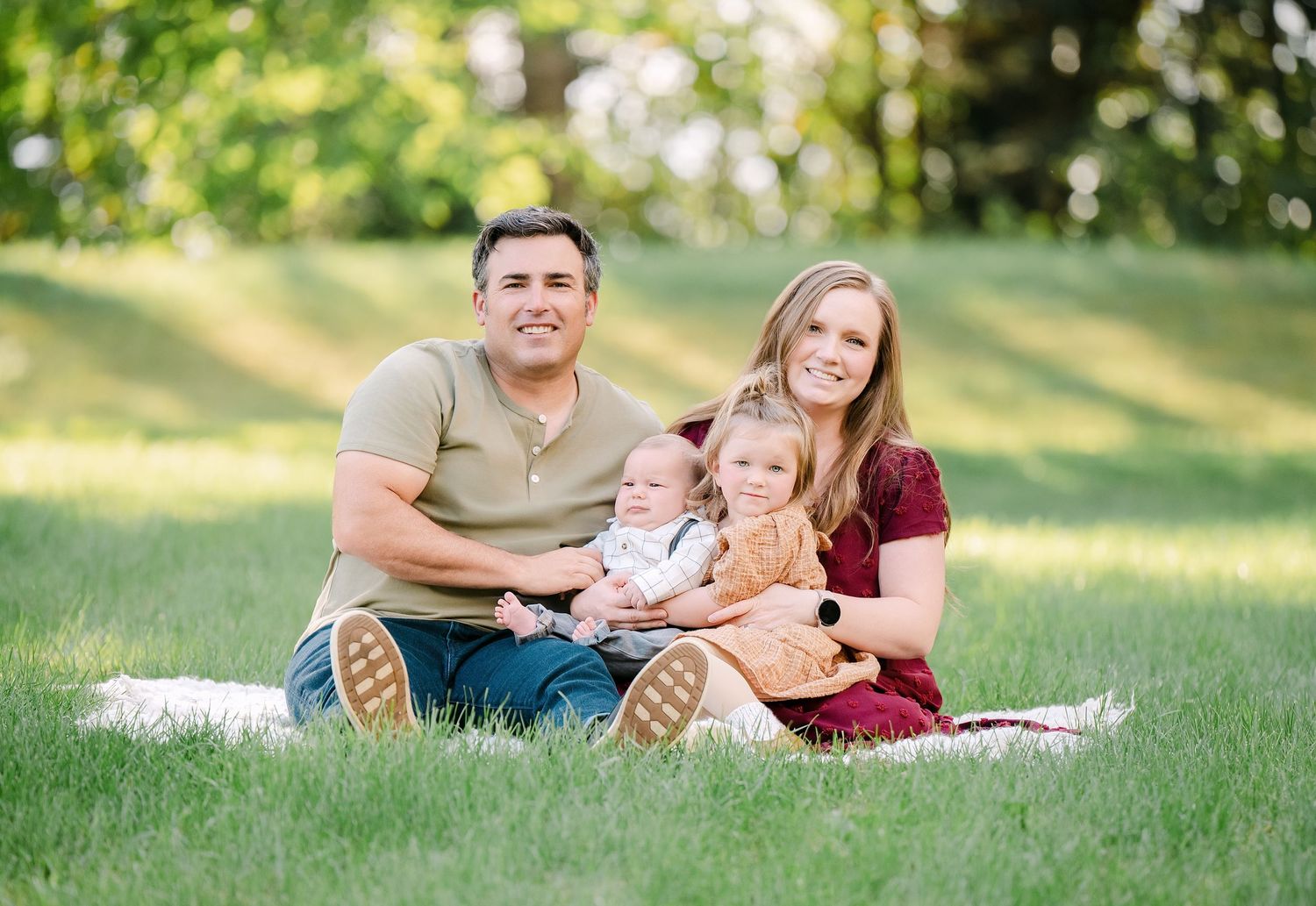 Happy family of four sitting together on grass outdoors during a sunny day portrait session.. Minnesota milestone photographer