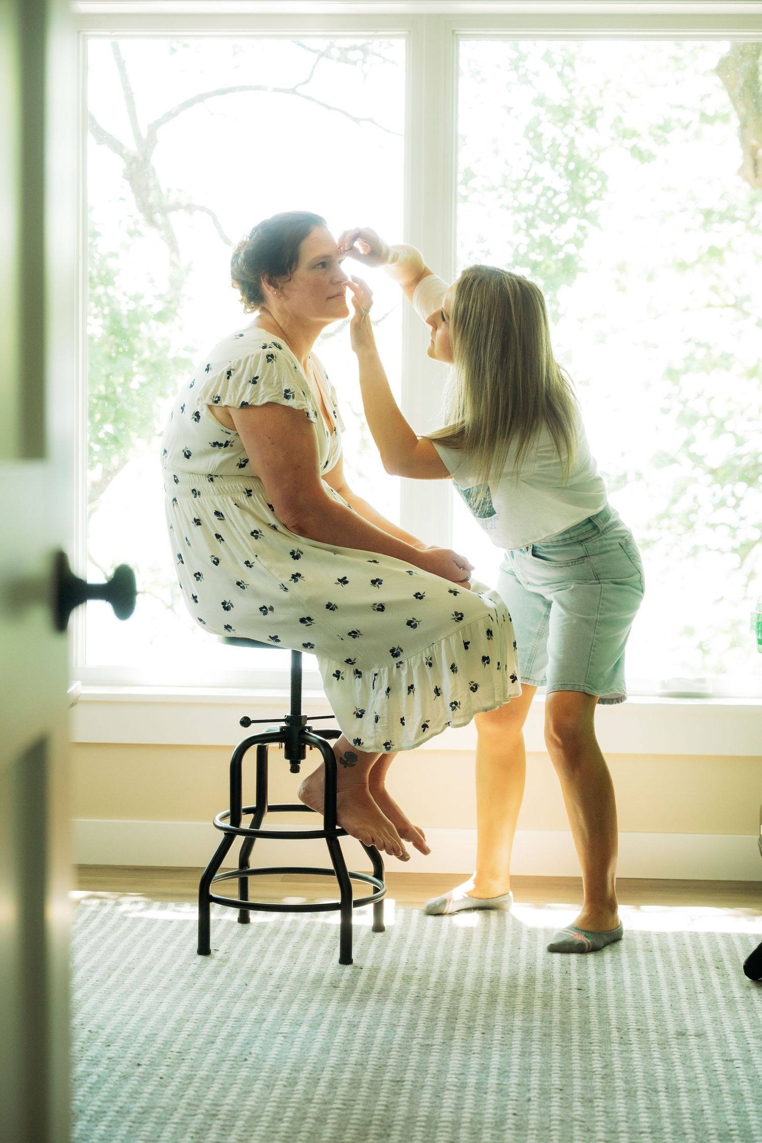 A young girl applies makeup to a woman sitting on a stool near a bright window in a cozy room.