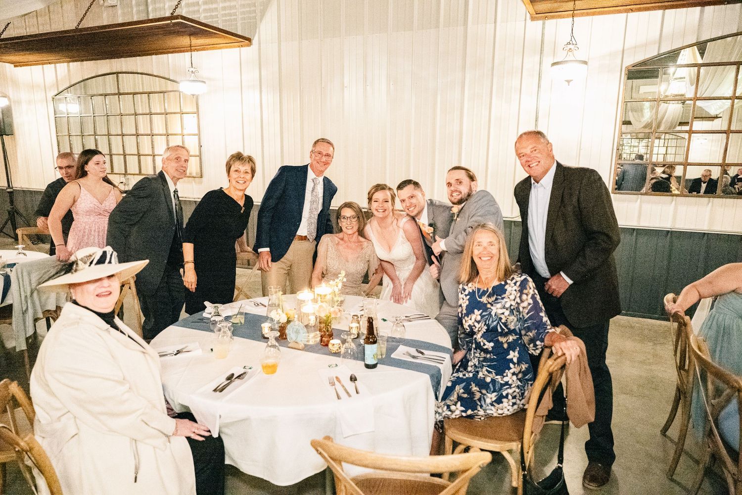 Wedding party and family gather for group photo at decorated reception dinner table.