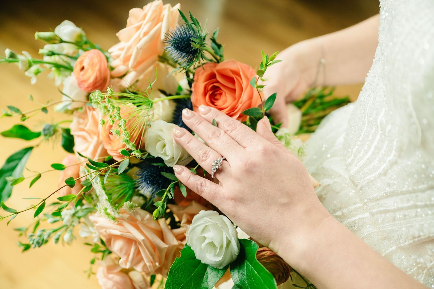 Bride's hand with diamond ring resting on a bouquet of peach roses, white flowers, and blue thistles.