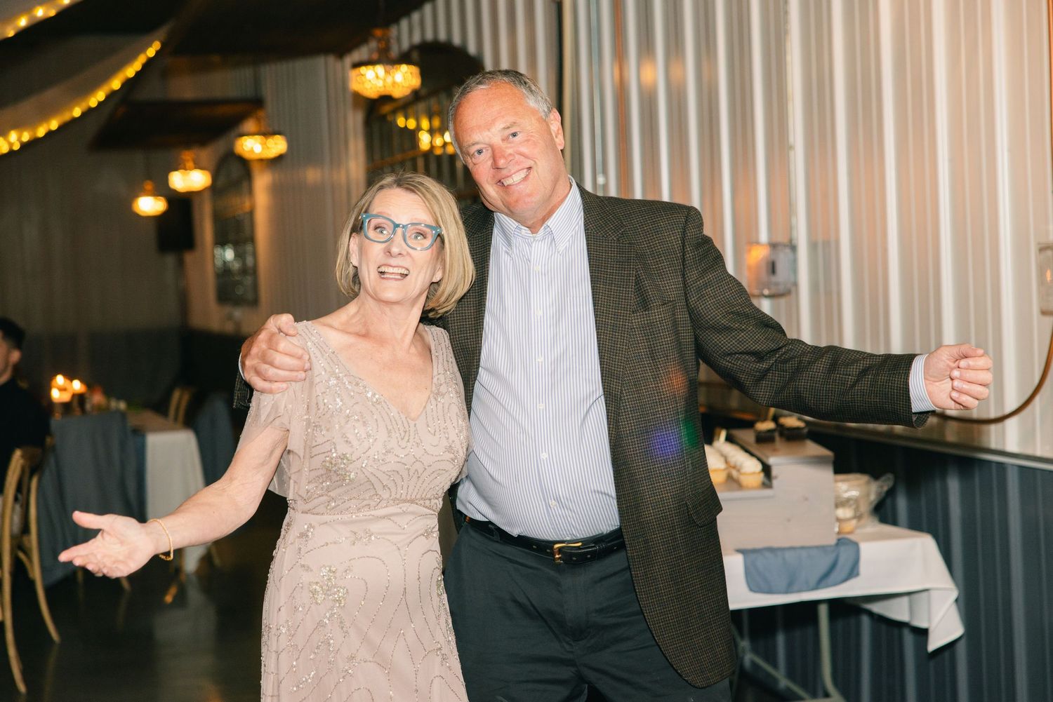 Smiling couple posing playfully with open arms at elegant wedding reception venue.