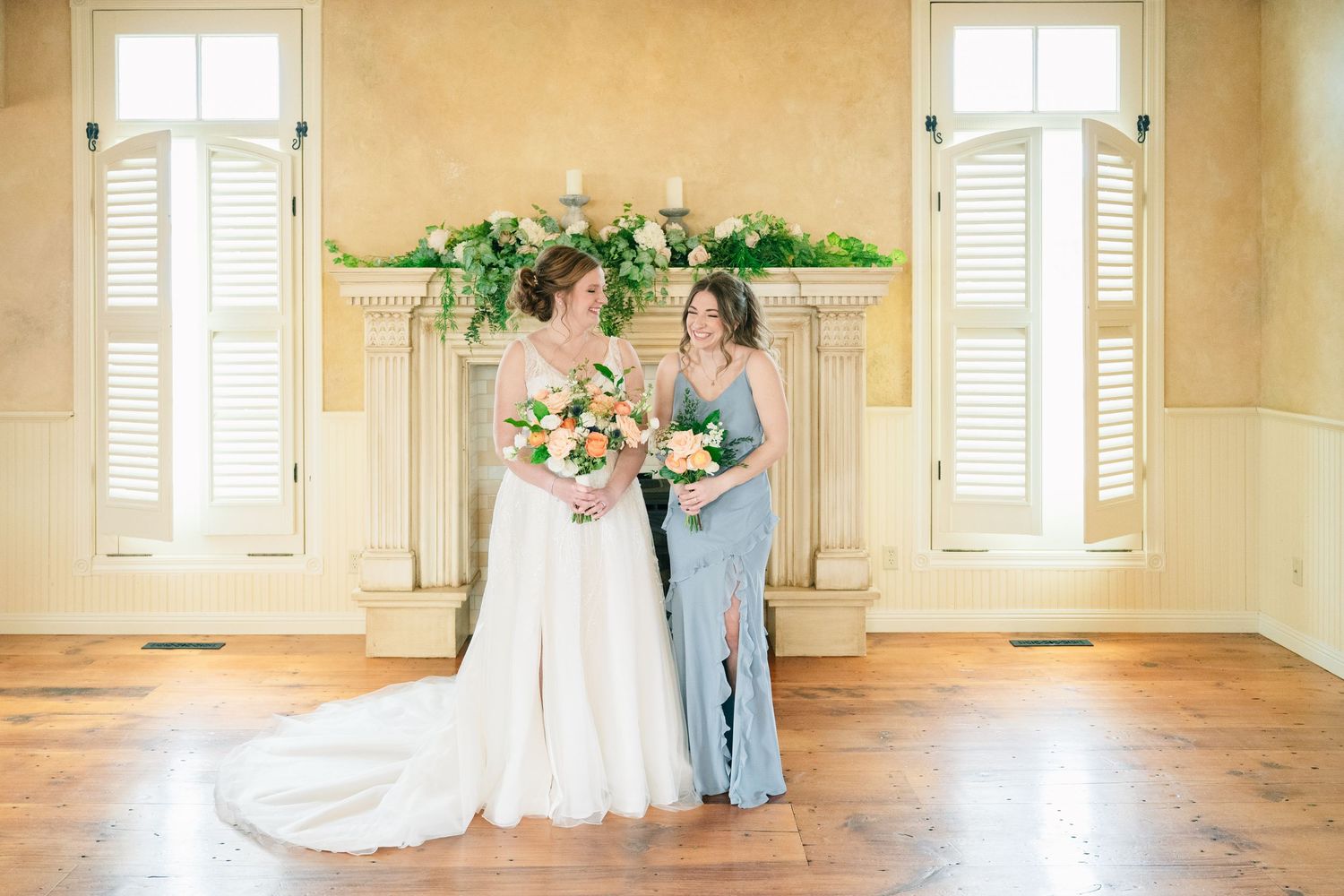 Bride in white gown and bridesmaid in blue dress holding bouquets by elegant fireplace with greenery.